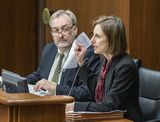 Becca Nash sits at the testifiers' table and speaks to someone out of view while gesturing with one hand. Representative Hansen sits next to her and looks at her while she speaks.